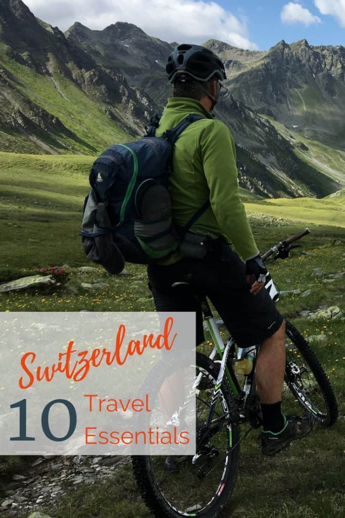 Image of man with backpack on bike riding though the Swiss Alps countryside with text overlay: "10 Switzerland Travel Essential".